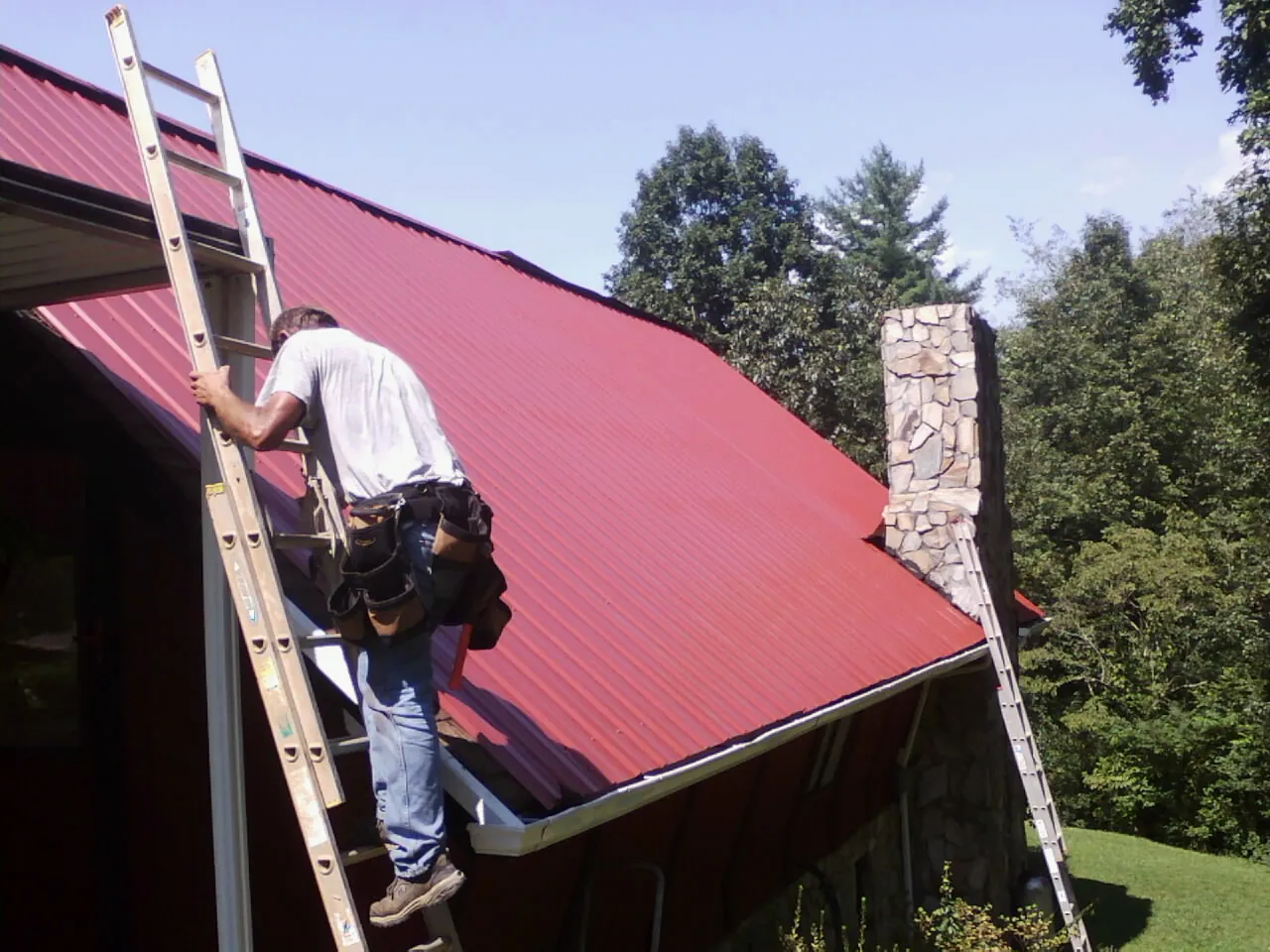 Roofer on a ladder beside a red metal roof and stone chimney — Soffit & Fascia Repair by Roofing Brothers in Monticello